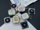 Clear earbud case filled with golden brown liquid glitter, attached to a gold cat-shaped clip and a miniature vinyl record keychain.