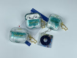 Clear earbud case filled with emerald green liquid glitter, attached to a gold cat-shaped clip and a miniature vinyl record keychain.