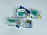 Clear earbud case filled with emerald green liquid glitter, attached to a gold cat-shaped clip and a miniature vinyl record keychain.
