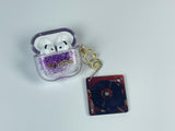 Clear earbud case filled with holographic lavender and blue liquid glitter, attached to a gold cat-shaped clip and a miniature vinyl record keychain.