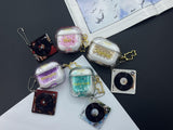 Clear earbud case filled with ghostly silver grayscale liquid glitter, attached to a gold cat-shaped clip and a miniature vinyl record keychain.
