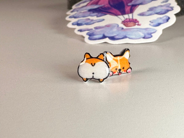 A pair of tiny resin stud earrings featuring a mismatched design of a happy corgi face and a corgi backside.