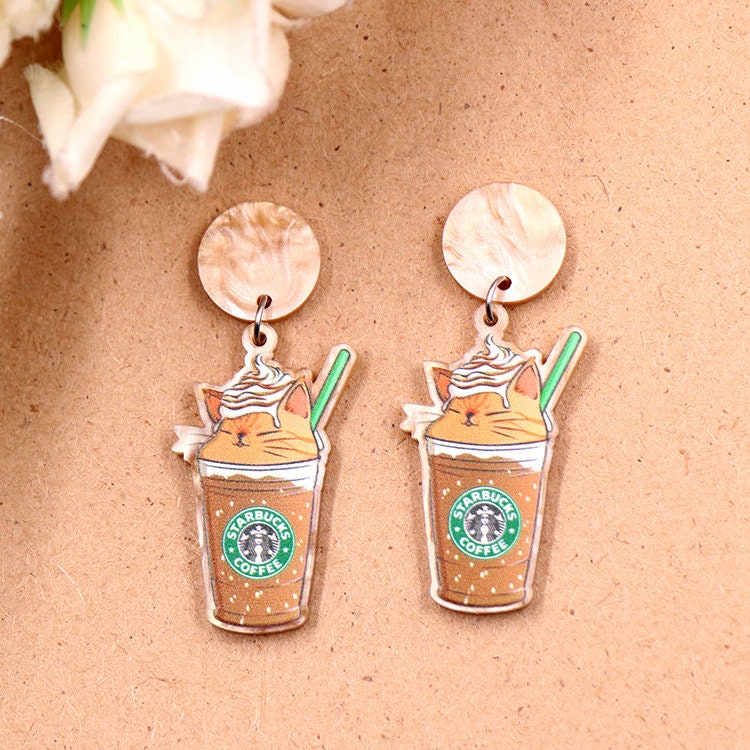 A pair of acrylic dangle earrings featuring a sleepy orange cat inside an iced coffee cup with whipped cream, hanging from marbled beige resin studs.