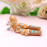 A pair of acrylic dangle earrings featuring a sleepy orange cat inside an iced coffee cup with whipped cream, hanging from marbled beige resin studs.
