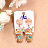A pair of acrylic dangle earrings featuring a sleepy orange cat inside an iced coffee cup with whipped cream, hanging from marbled beige resin studs.